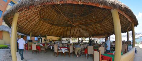 An outdoor dining area with thatched roof and multiple tables and chairs.