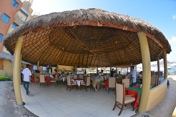 Un área de comedor al aire libre con techo de paja y varias mesas y sillas.
