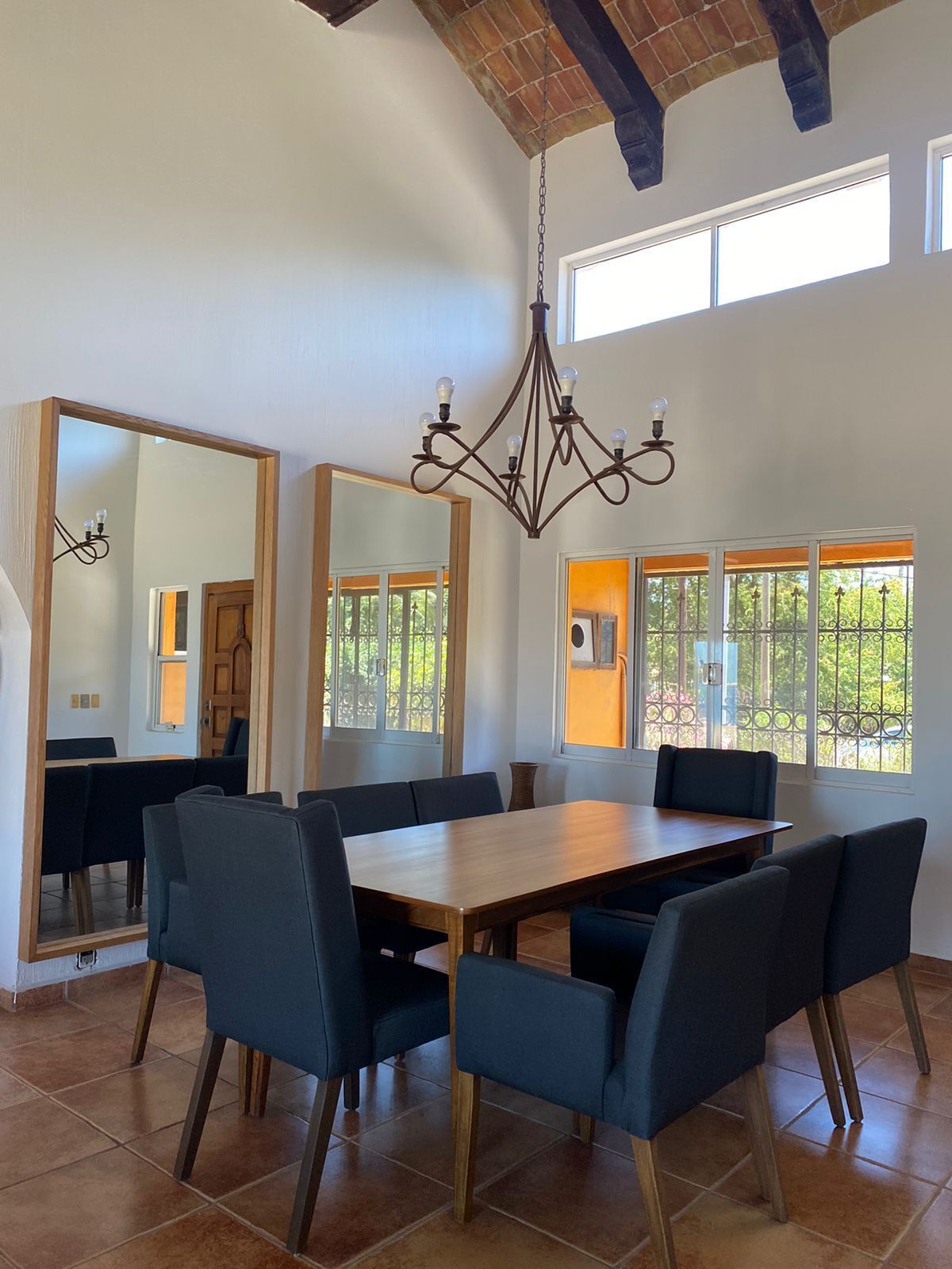 A dining area with a large wooden table, dark blue chairs, a chandelier, and large windows with views of greenery.