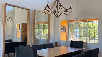 A dining area with a large wooden table, dark blue chairs, a chandelier, and large windows with views of greenery.
