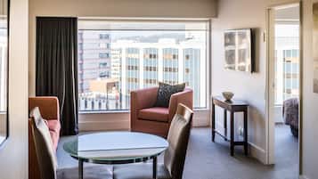 A hotel room with a glass table, two chairs, a sofa, a small side table, a framed picture, and a view of buildings through a large window.