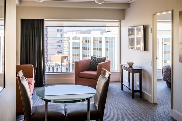 A hotel room with a glass table, two chairs, a sofa, a small side table, a framed picture, and a view of buildings through a large window.