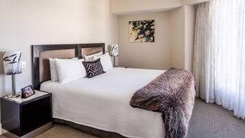 A neatly made bed with a white comforter and a fur throw, a bedside table with a lamp and a tablet, and a painting on the wall.