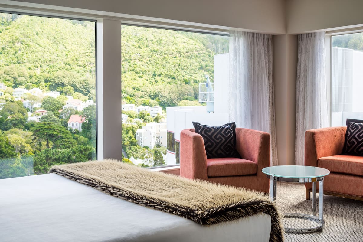 Ein Hotelzimmer mit einem großen Bett, zwei Sesseln, einem kleinen Glastisch und Blick auf eine Hügellandschaft mit Gebäuden.
