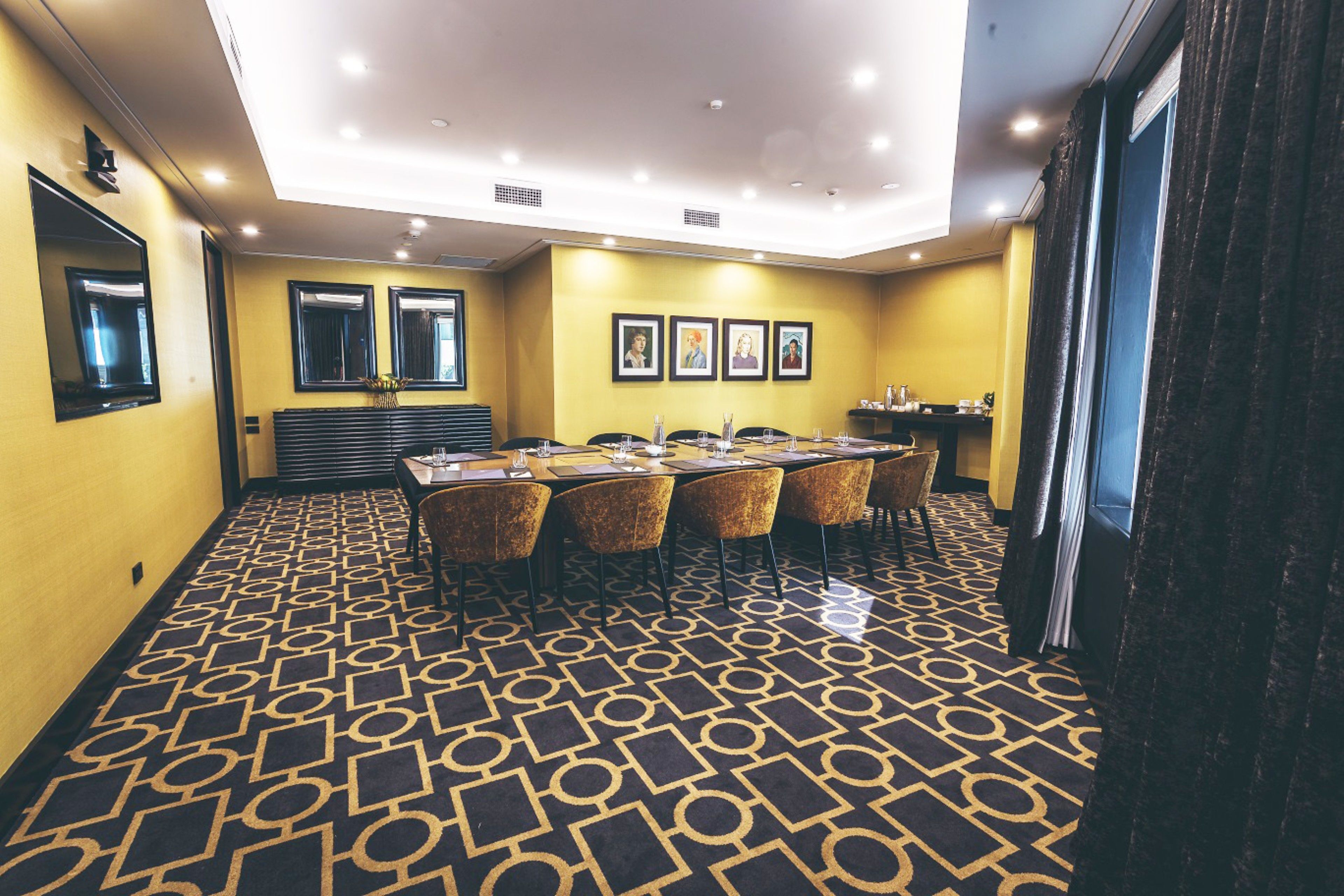 Una sala de conferències amb un mosaic de moqueta, una taula llarga amb cadires, quadres als murs i una finestra amb cortines.