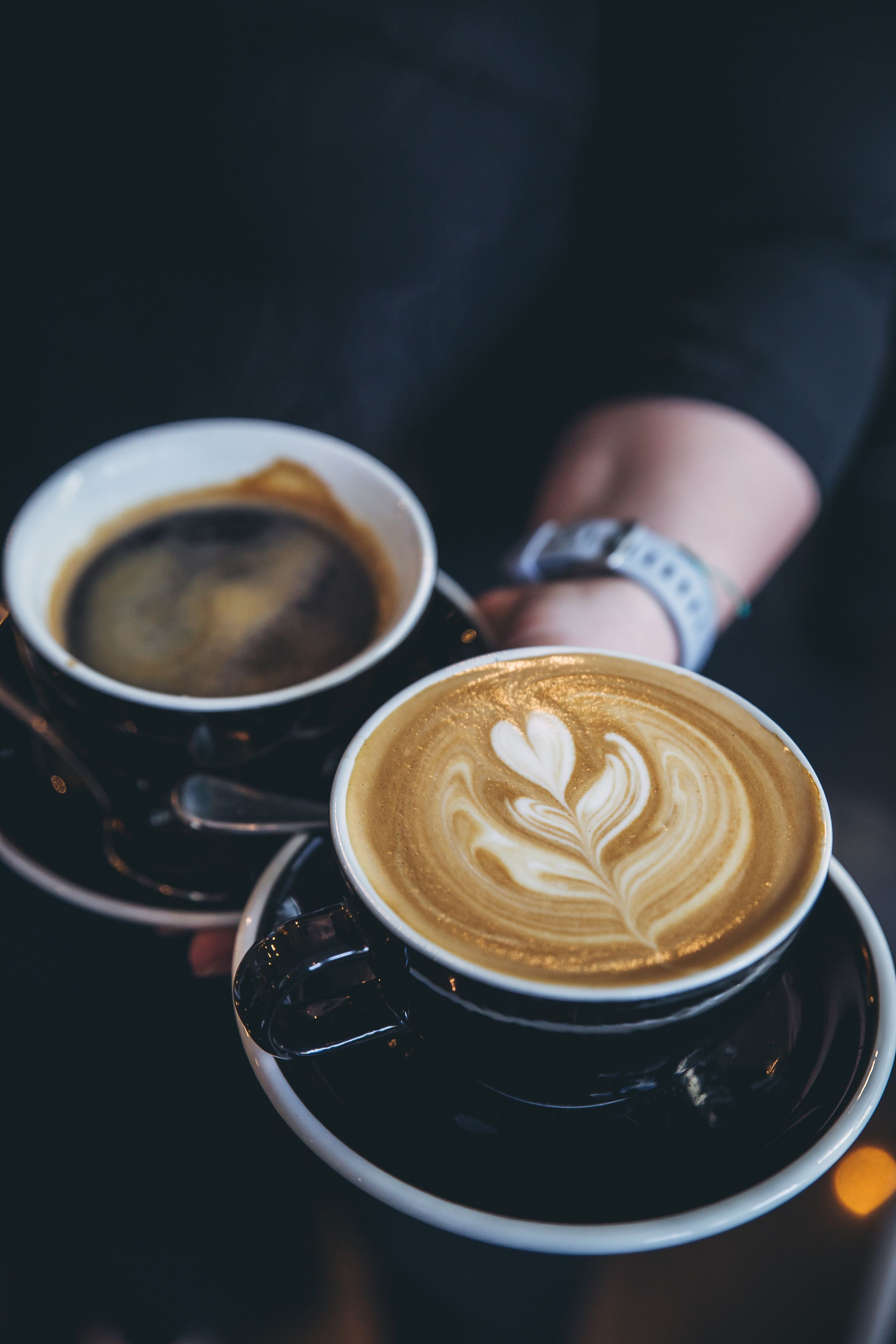 Zwei Tassen Kaffee mit Latte-Art, gehalten von einer Person, die eine Uhr trägt.