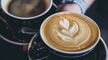 Zwei Tassen Kaffee mit Latte-Art, gehalten von einer Person, die eine Uhr trägt.