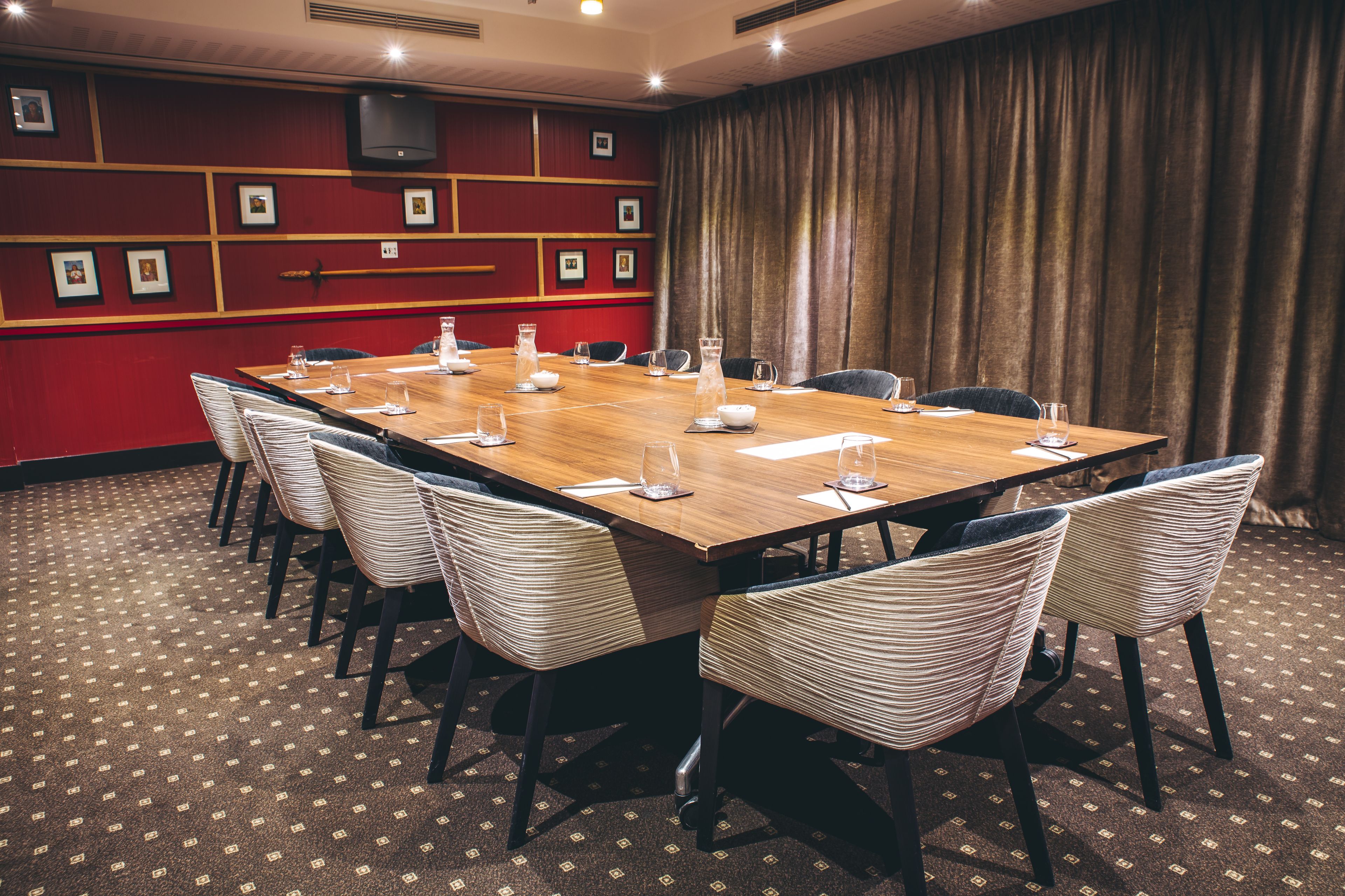 Une salle de conférence avec une longue table en bois, des chaises blanches recouvertes de tissu et un mur rouge orné de tableaux.