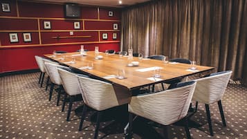 Une salle de conférence avec une longue table en bois, des chaises blanches recouvertes de tissu et un mur rouge orné de tableaux.