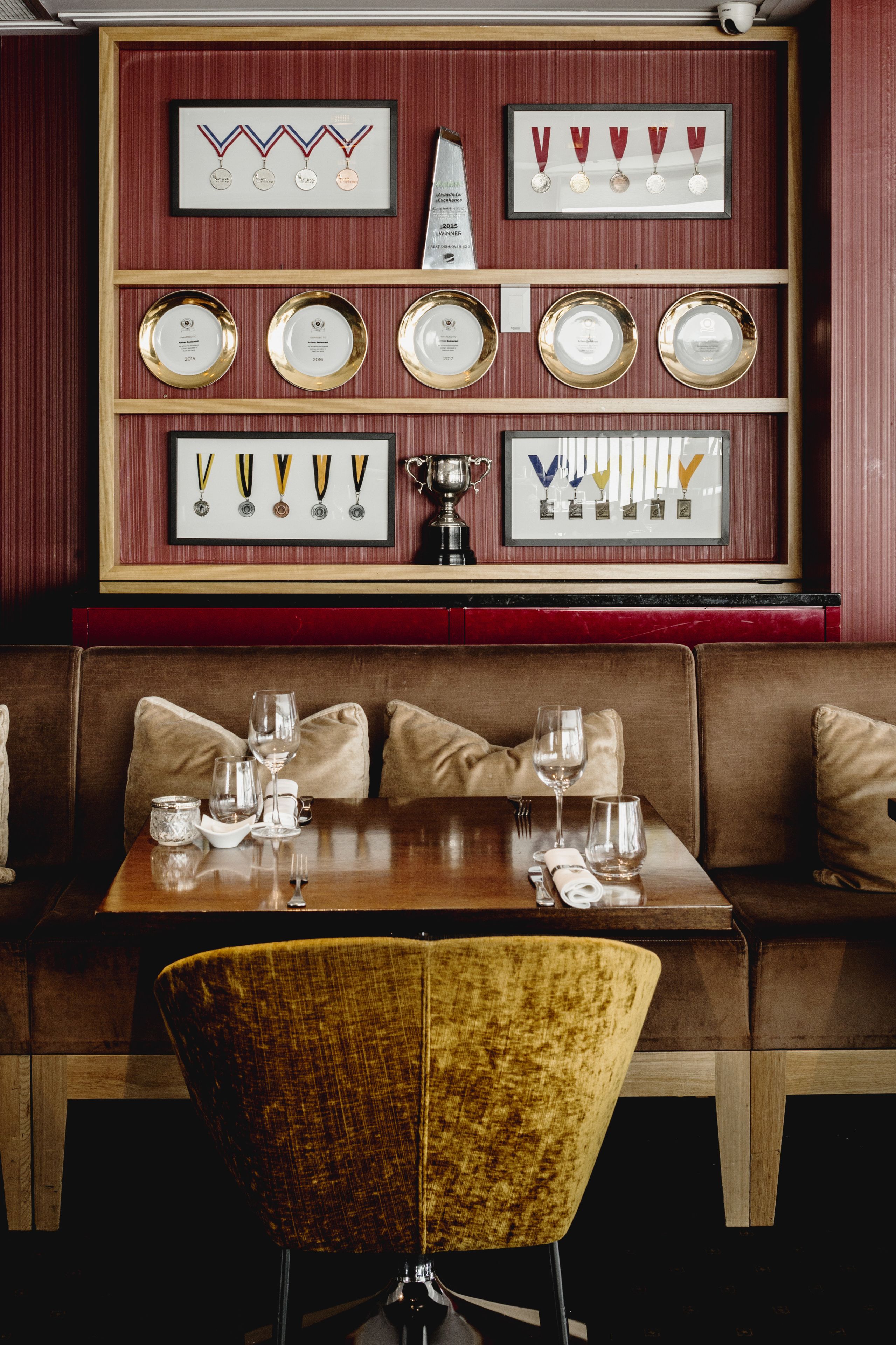 L’intérieur d’un restaurant présente une table en bois dressée pour le repas, un banc rembourré et un mur où sont exposés, encadrés, des prix et des médailles.