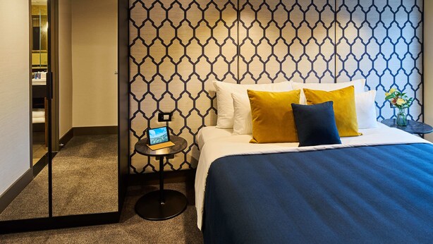 Quarto de hotel com cama, mesa de cabeceira, espelho e parede com padrão geométrico.