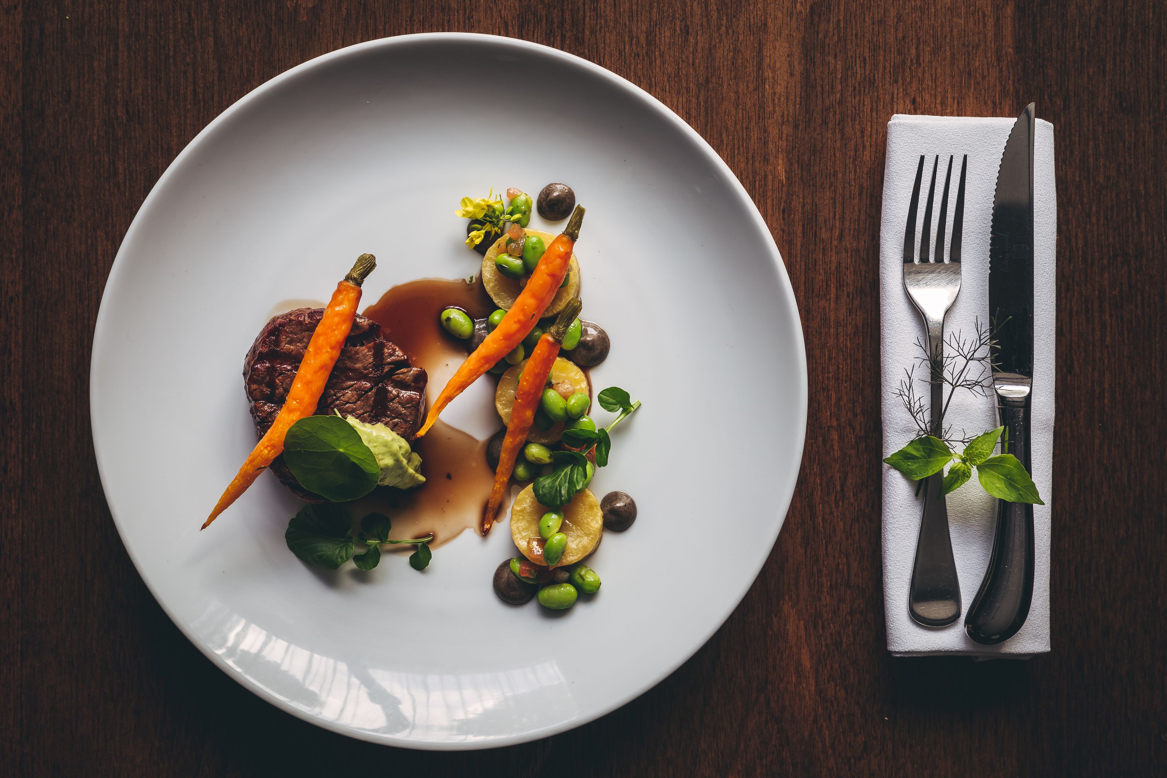 A plate of food with a steak, carrots, peas, and greens, accompanied by a fork and knife on a napkin.