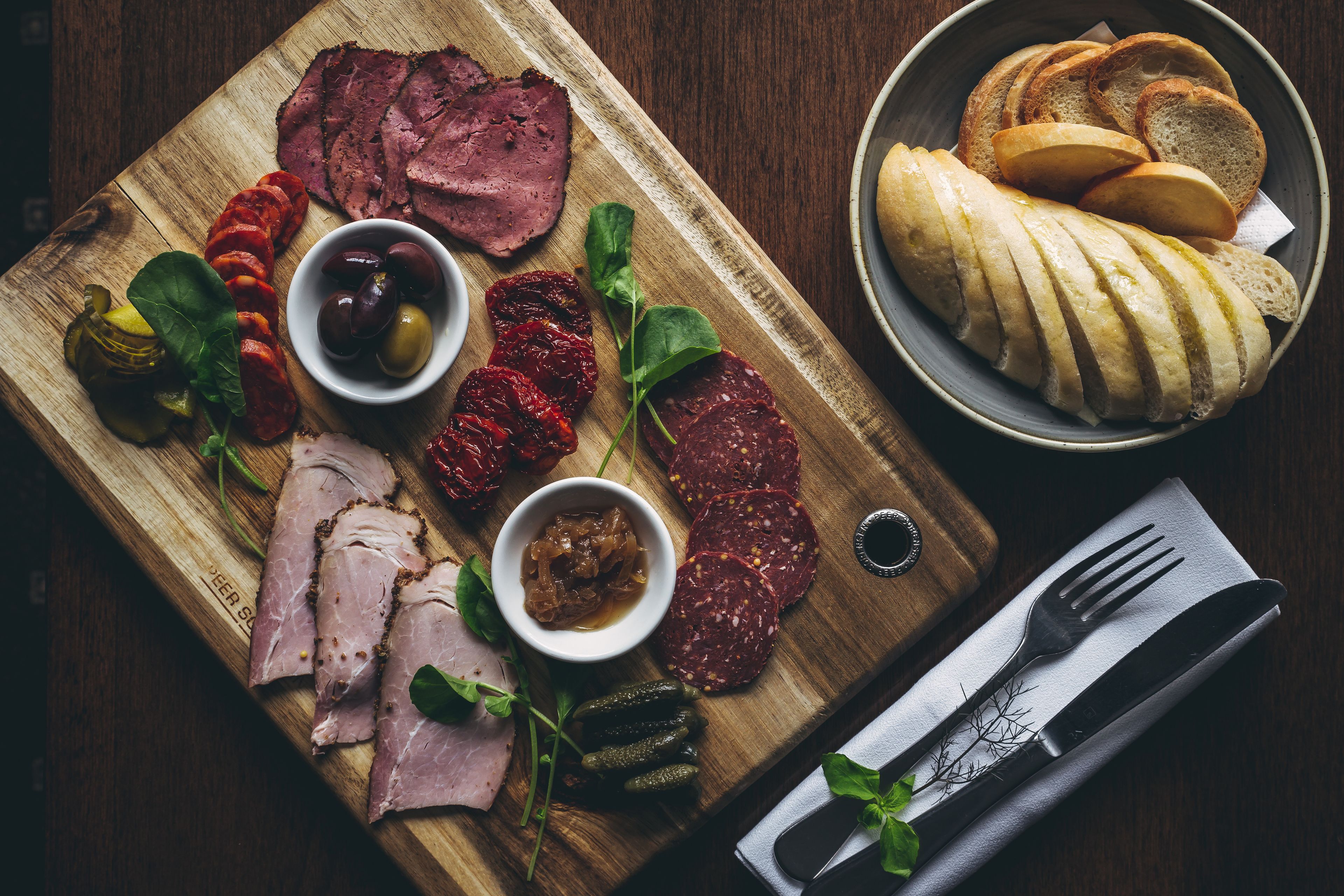 A wooden board with sliced meats, pickles, olives, and bread.