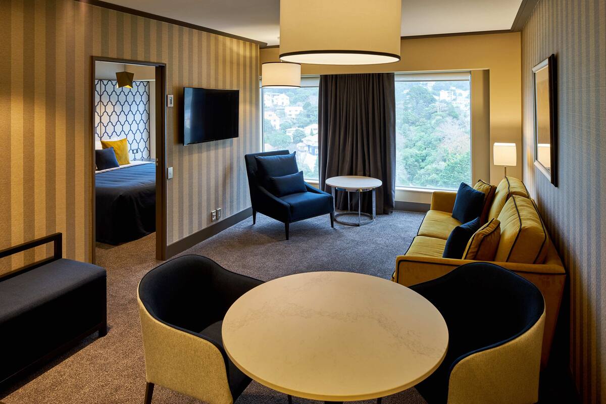 Ein Hotelzimmer mit Bett, Sofa, Sessel, rundem Tisch, Fernseher und Fenster mit Blick auf Gebäude.