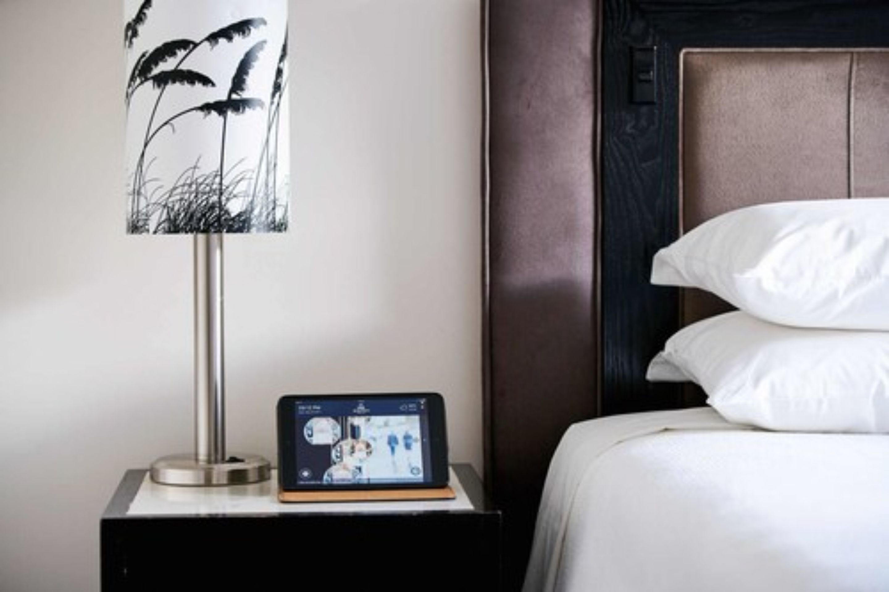 Un letto rifatto con lenzuola e cuscini bianchi, un comodino con una lampada che mostra un disegno floreale in bianco e nero, e un tablet sul tavolo.