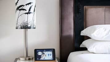 Un letto rifatto con lenzuola e cuscini bianchi, un comodino con una lampada che mostra un disegno floreale in bianco e nero, e un tablet sul tavolo.