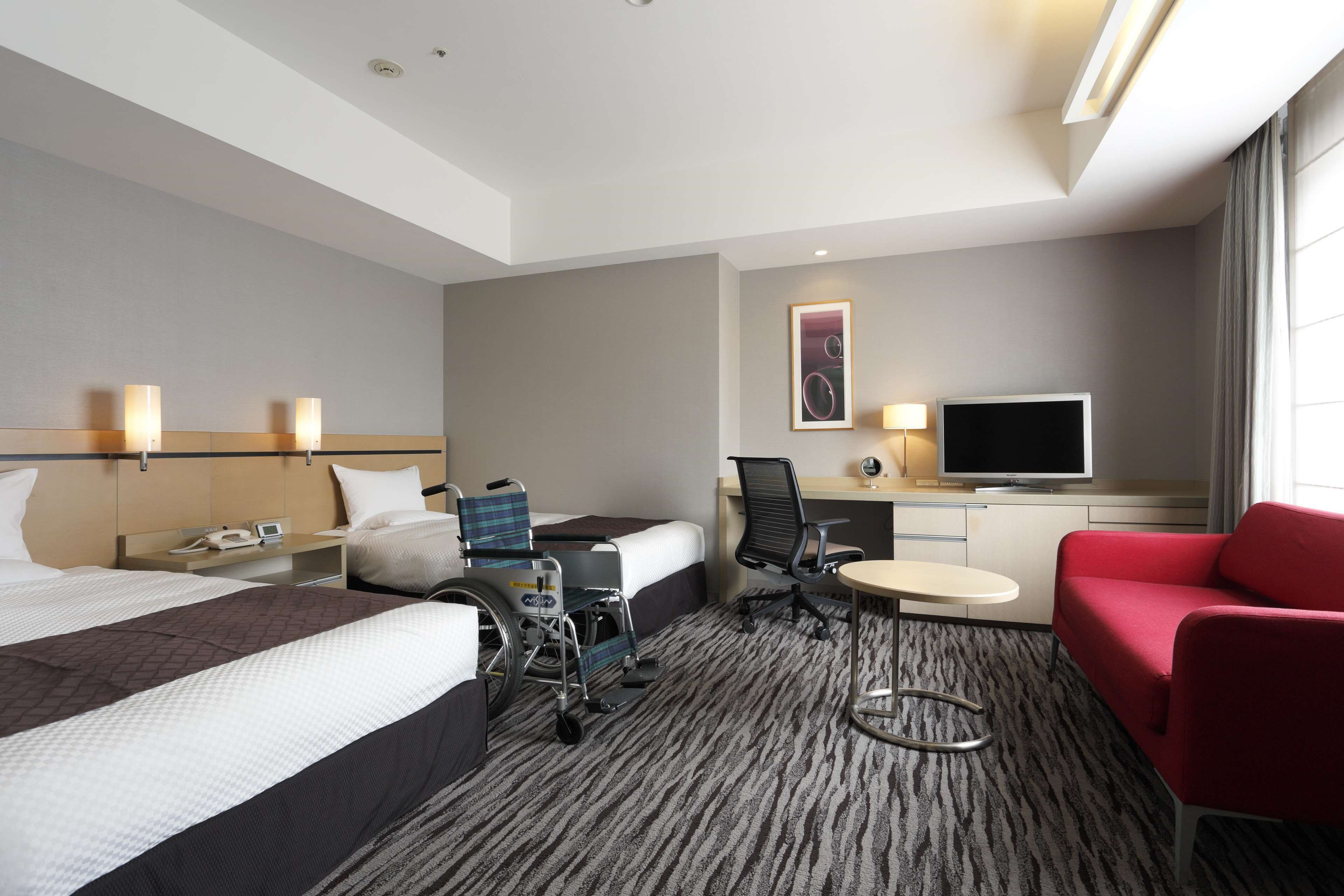 A hotel room with a bed, a desk with a TV, a wheelchair, a red chair, and a small table.