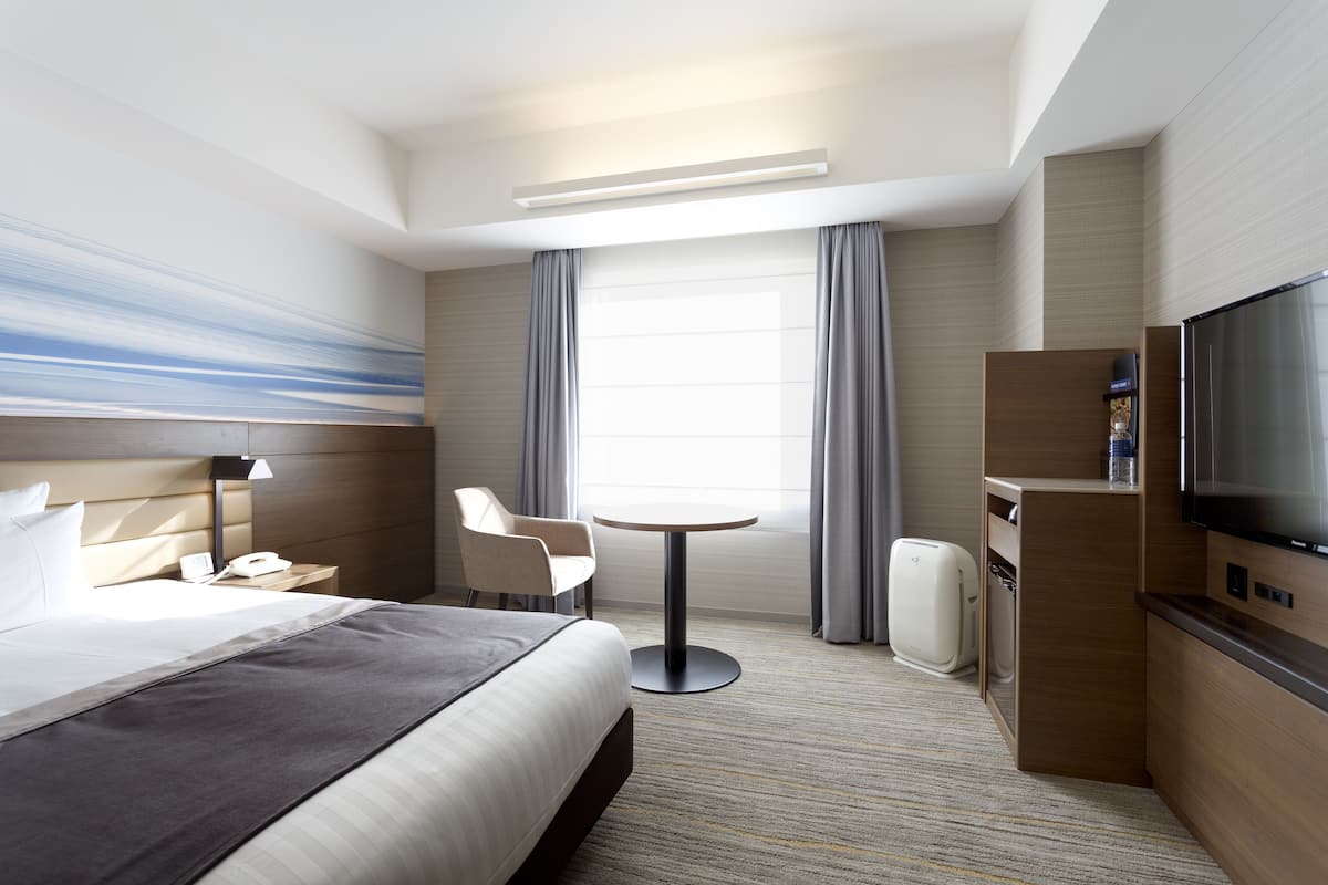 A hotel room with a bed, a desk, a chair, a small table, a television, and a window with curtains.