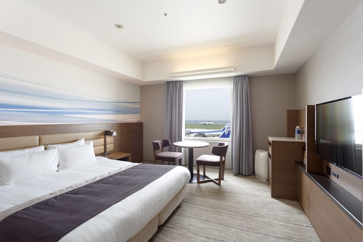 A hotel room with a large bed, a desk with a flat-screen TV, a small table, two chairs, and a window with a view of an airport.