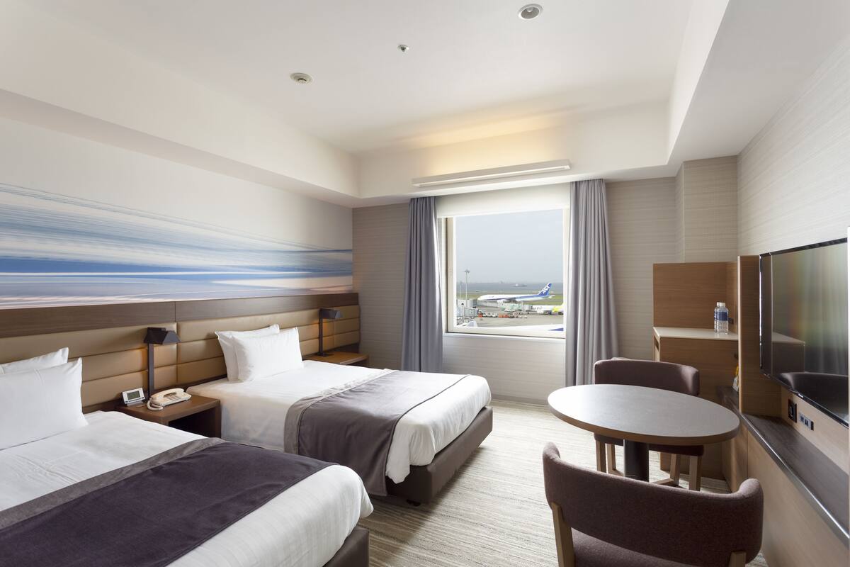 A hotel room with two beds, a desk, a chair, a TV, and a window with a view of an airport.