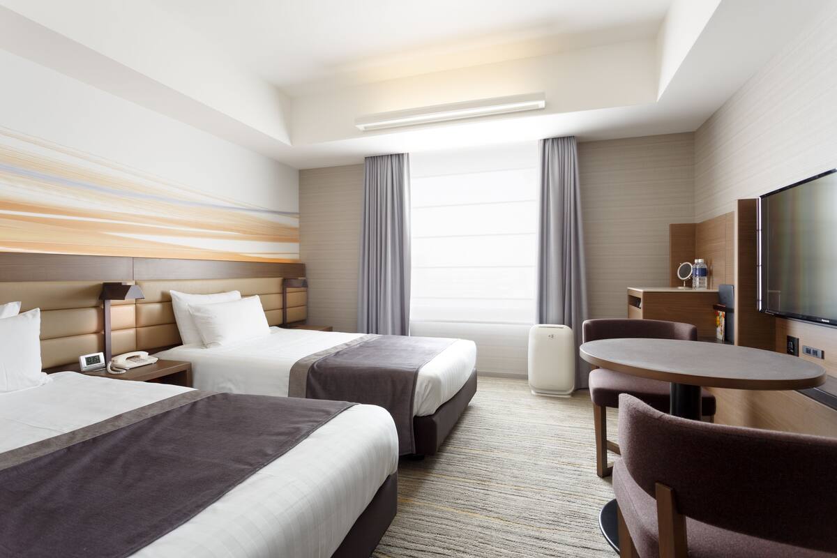 A hotel room with two beds, a desk, a chair, a television, and a large window with curtains.