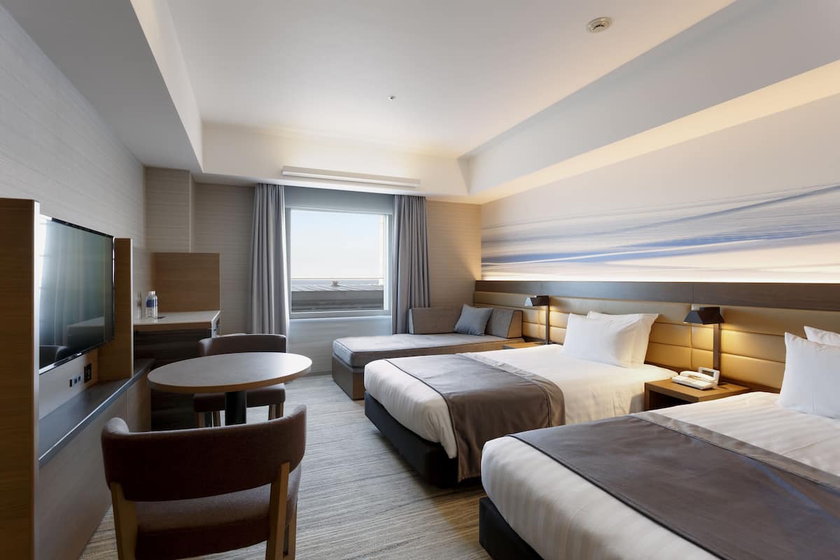 A hotel room with two beds, a TV, a desk, and a view of the sea.
