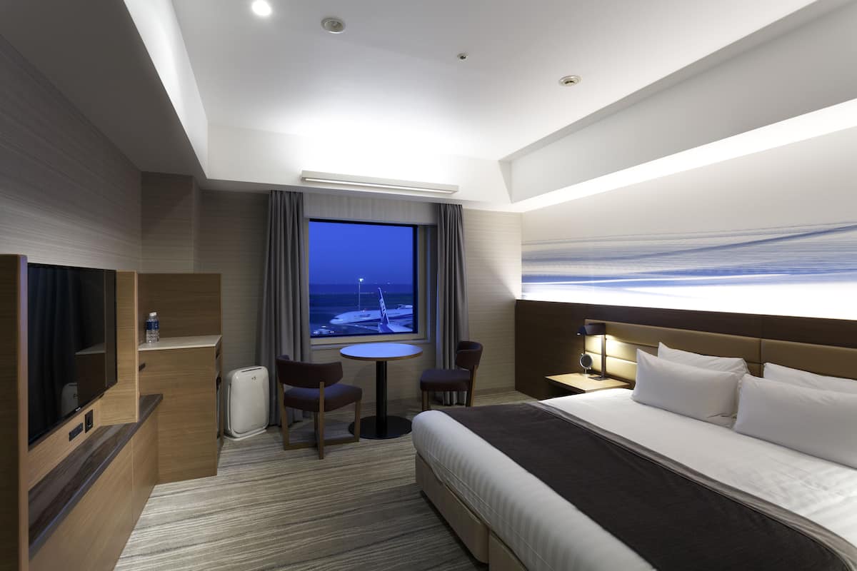 A hotel room with a bed, a TV, a desk, and a window with a view of the sea.