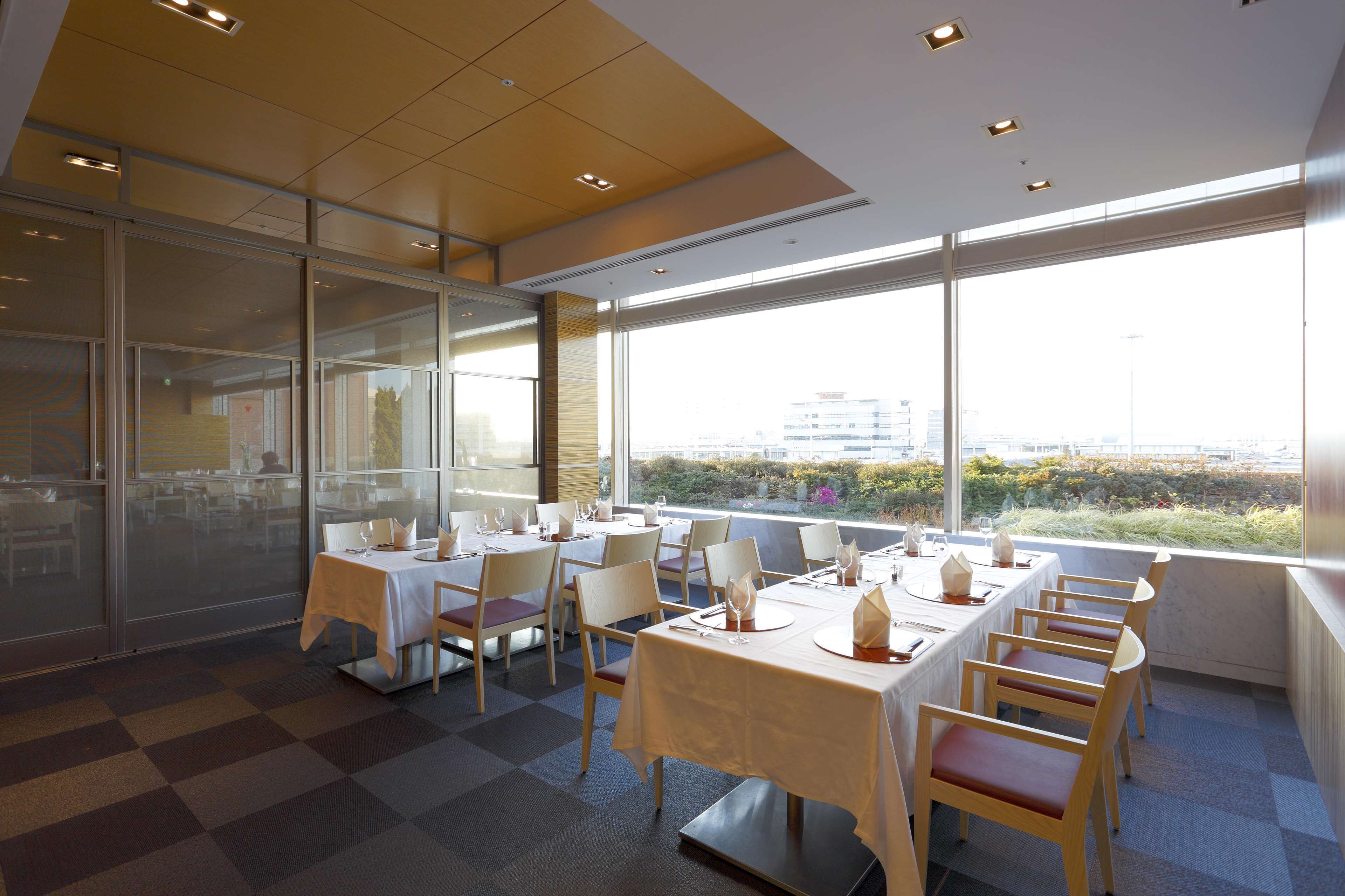Un espace repas avec des tables dressées pour le dîner, de grandes fenêtres offrant une vue sur la ville et un motif à carreaux sur le sol.