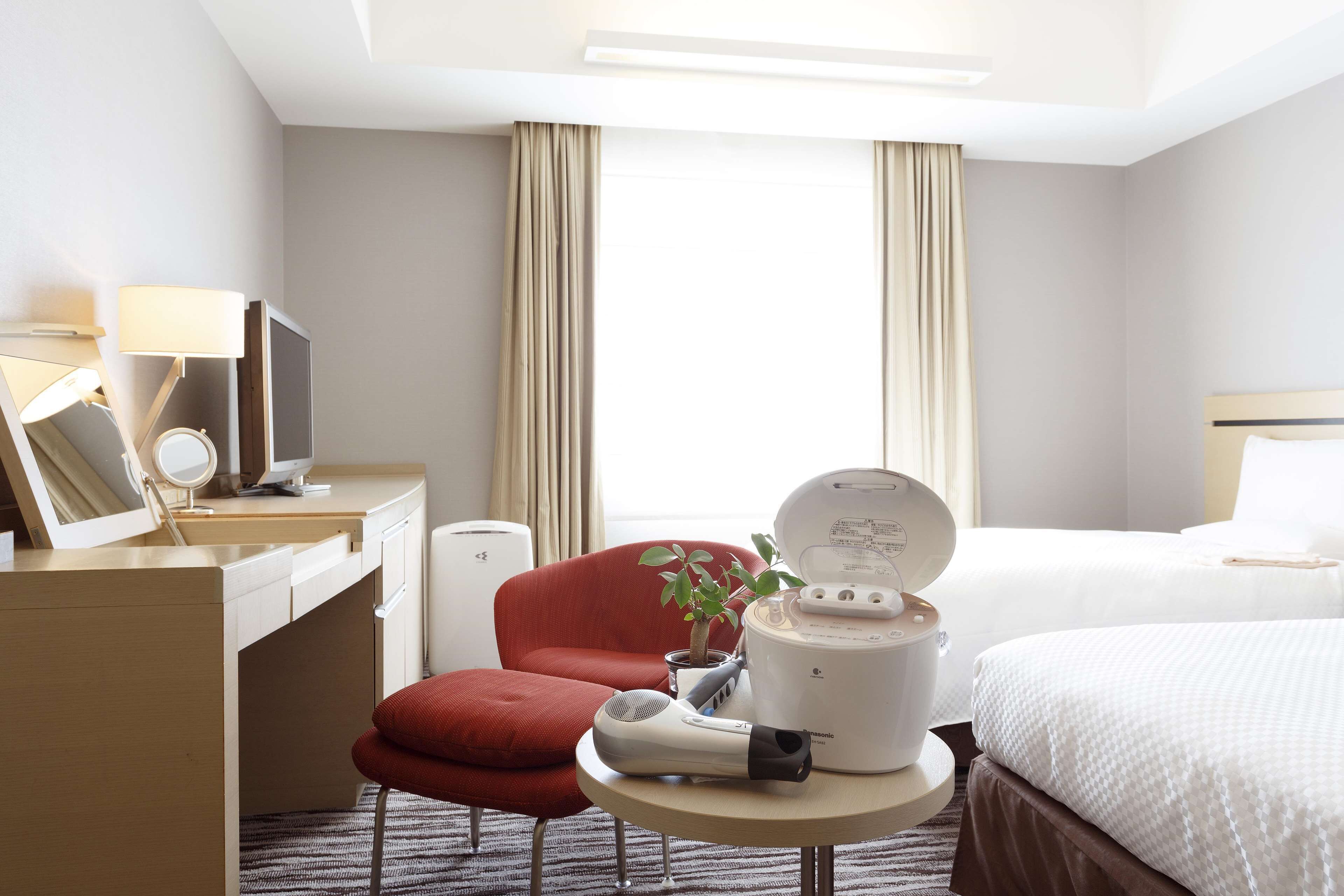 A hotel room with a desk, a red chair, a bed, a bedside table with a lamp, a hairdryer, and a plant.