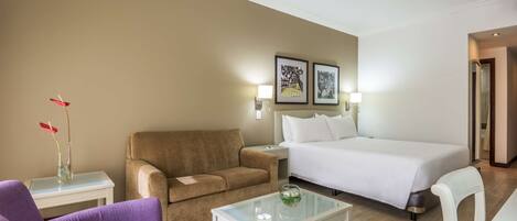 A hotel room with a bed, a sofa, a chair, a coffee table, and a nightstand with a lamp.