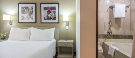 A hotel room with a bed, a nightstand, a framed picture, a small plant, and a bathroom with a bathtub and shower.