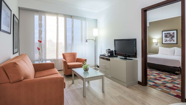 A modern hotel room with a sofa, a coffee table, a TV, a bed, and a view of the city through large windows.