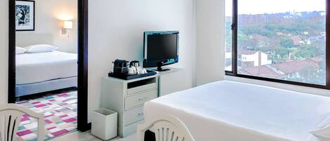 A hotel room with a bed, a desk with a computer and a TV, a chair, and a window with a city view.