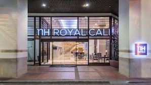 The entrance of NH Royal Cali hotel with a modern glass facade and a visible staircase leading inside.