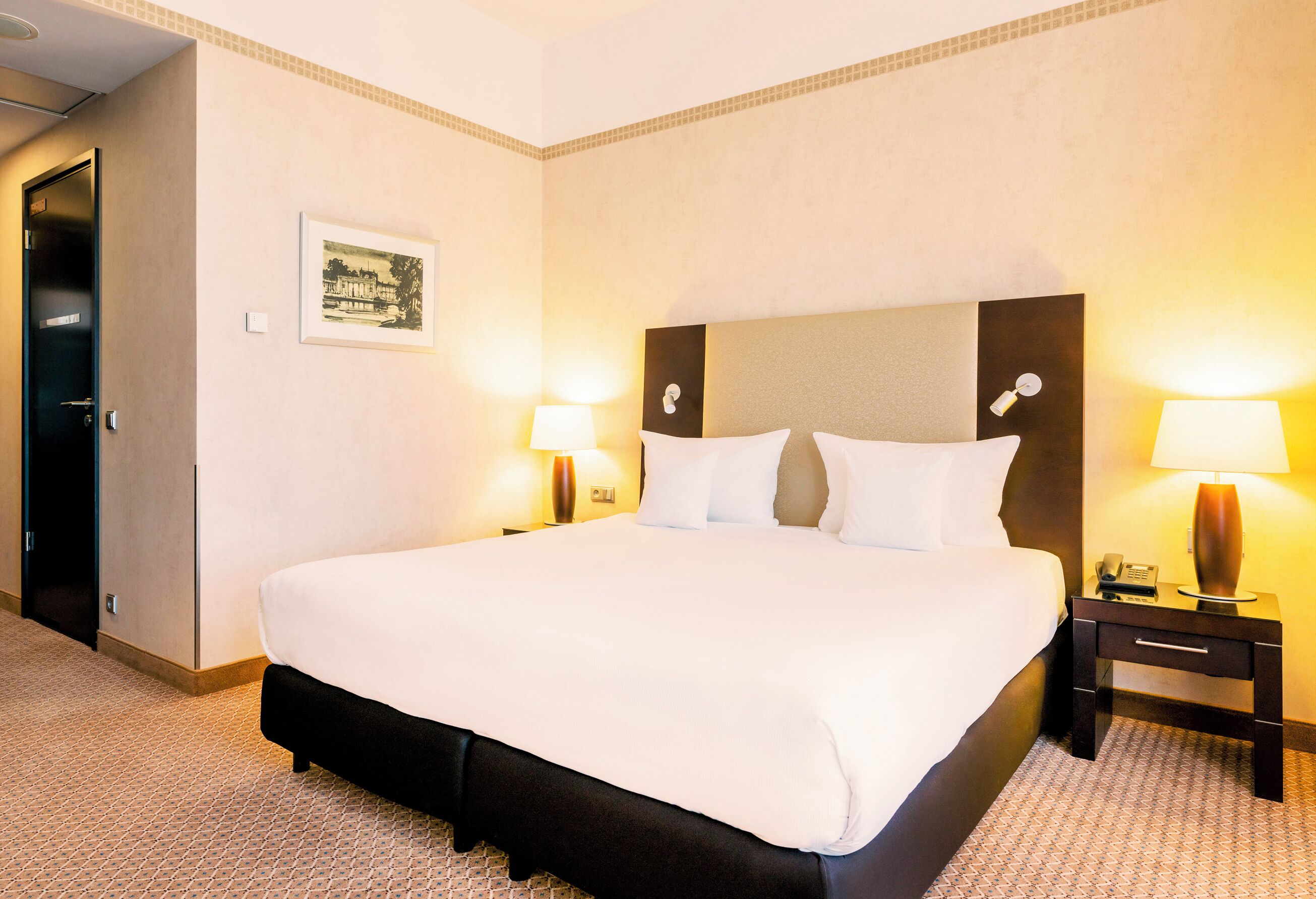 Een hotelkamer met een groot bed, twee nachtkastjes met lampen, een ingelijst schilderij aan de muur en een deur aan de linkerkant.