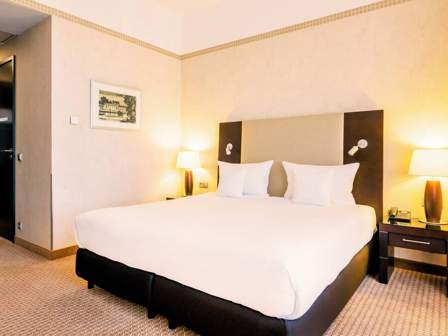 A hotel room with a large bed, two bedside tables with lamps, a framed picture on the wall, and a door to the left.