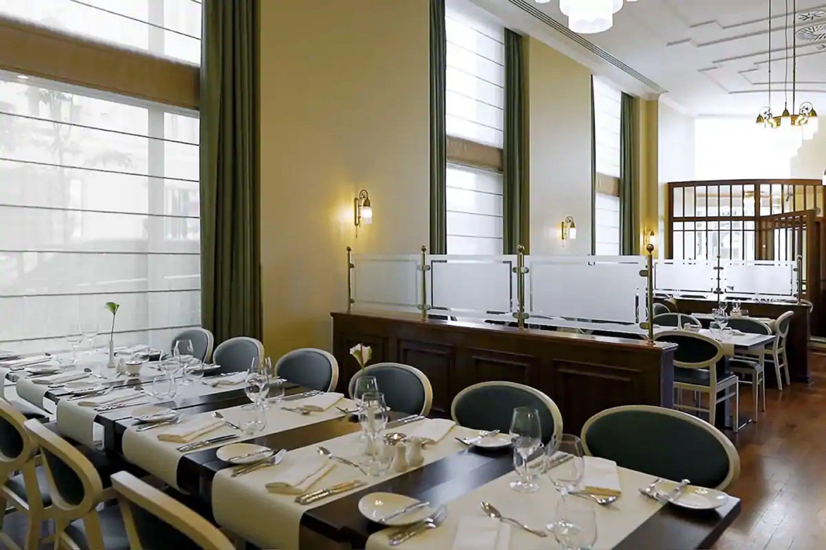 A dining area with multiple tables set for a meal, large windows with blinds, and a chandelier.