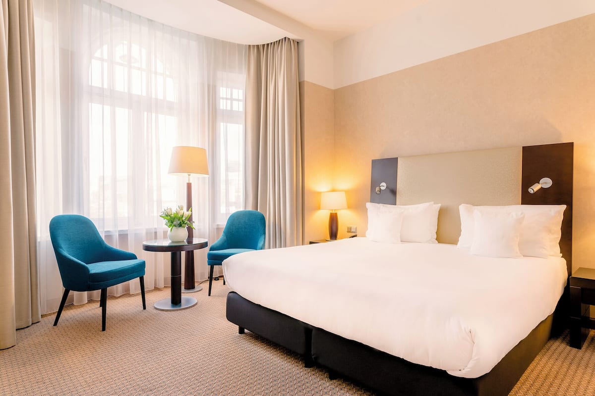 A hotel room with a large bed, two chairs, a small table with a lamp, and a potted plant.