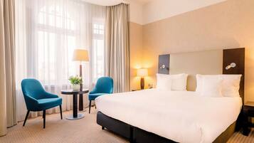 A hotel room with a large bed, two chairs, a small table with a lamp, and a potted plant.
