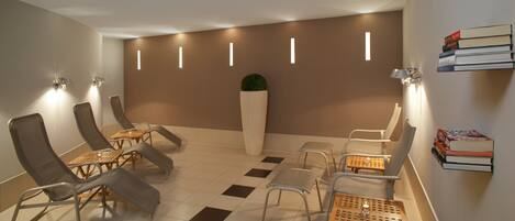 A modern lounge area with reclining chairs, a central planter, and a wall-mounted book shelf.