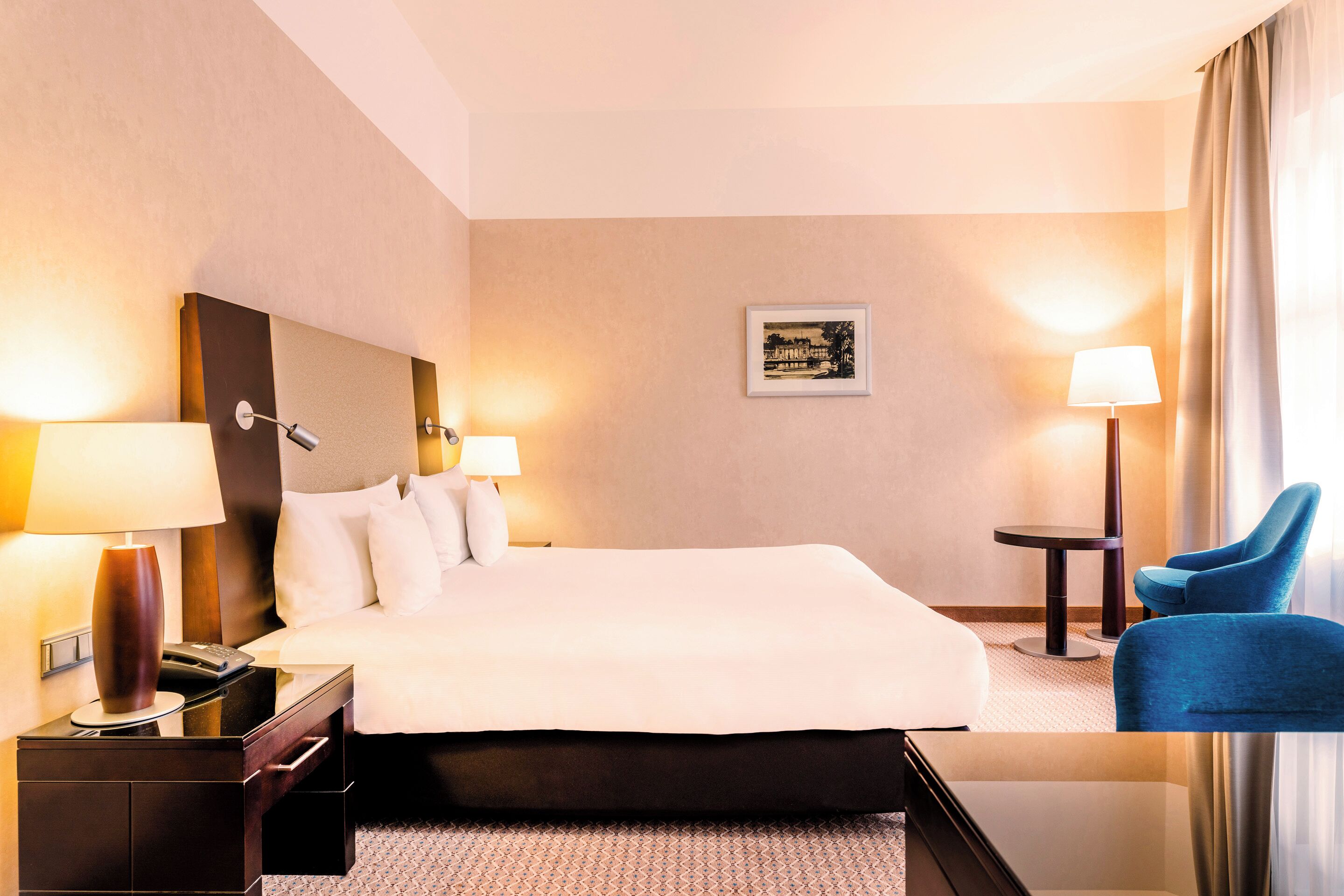 A hotel room with a large bed, bedside lamps, a small table, a chair, and a framed picture on the wall.