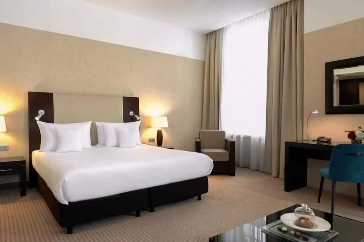 A hotel room with a large bed, bedside tables with lamps, a desk, a chair, and a view of the outdoors.