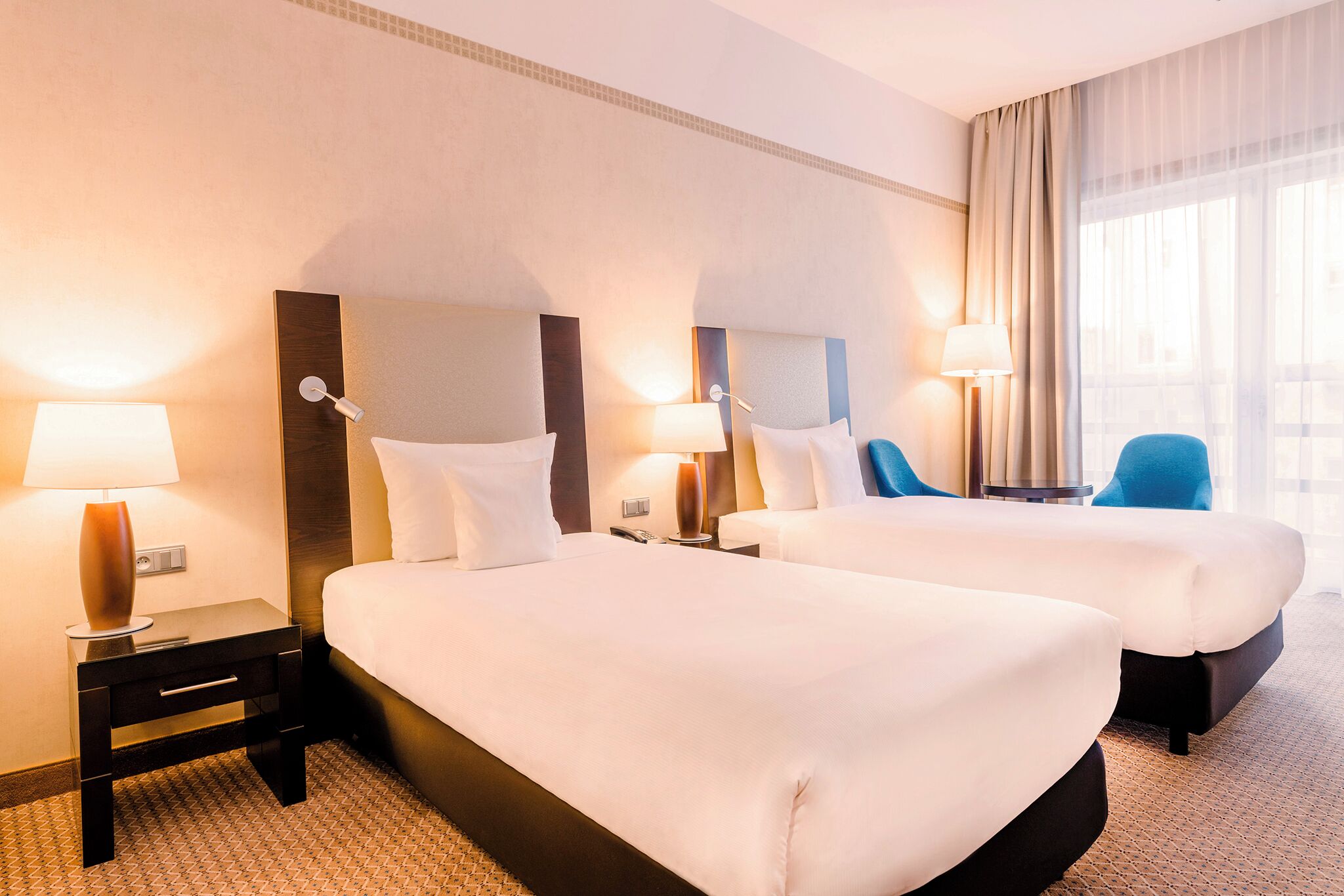 A hotel room with two beds, nightstands with lamps, a chair, and a large window with curtains.