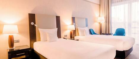 Habitación de hotel con dos camas, mesitas de noche con lámparas, una silla y un ventanal grande con cortinas.