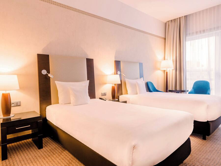 A hotel room with two beds, nightstands with lamps, a chair, and a large window with curtains.