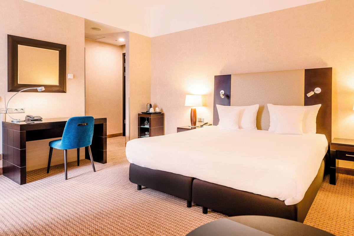A hotel room with a large bed, a desk, a chair, a desk lamp, a telephone, and a nightstand with a lamp.