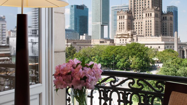 A balcony with a table, a vase of flowers, and a view of a cityscape including a prominent building with a clock tower. - Polonia Palace Hotel (Warsaw)