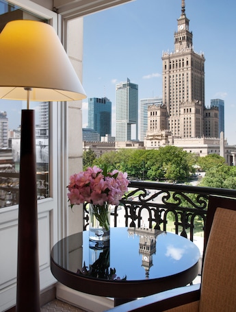 A balcony with a table, a vase of flowers, and a view of a cityscape including a prominent building with a clock tower.