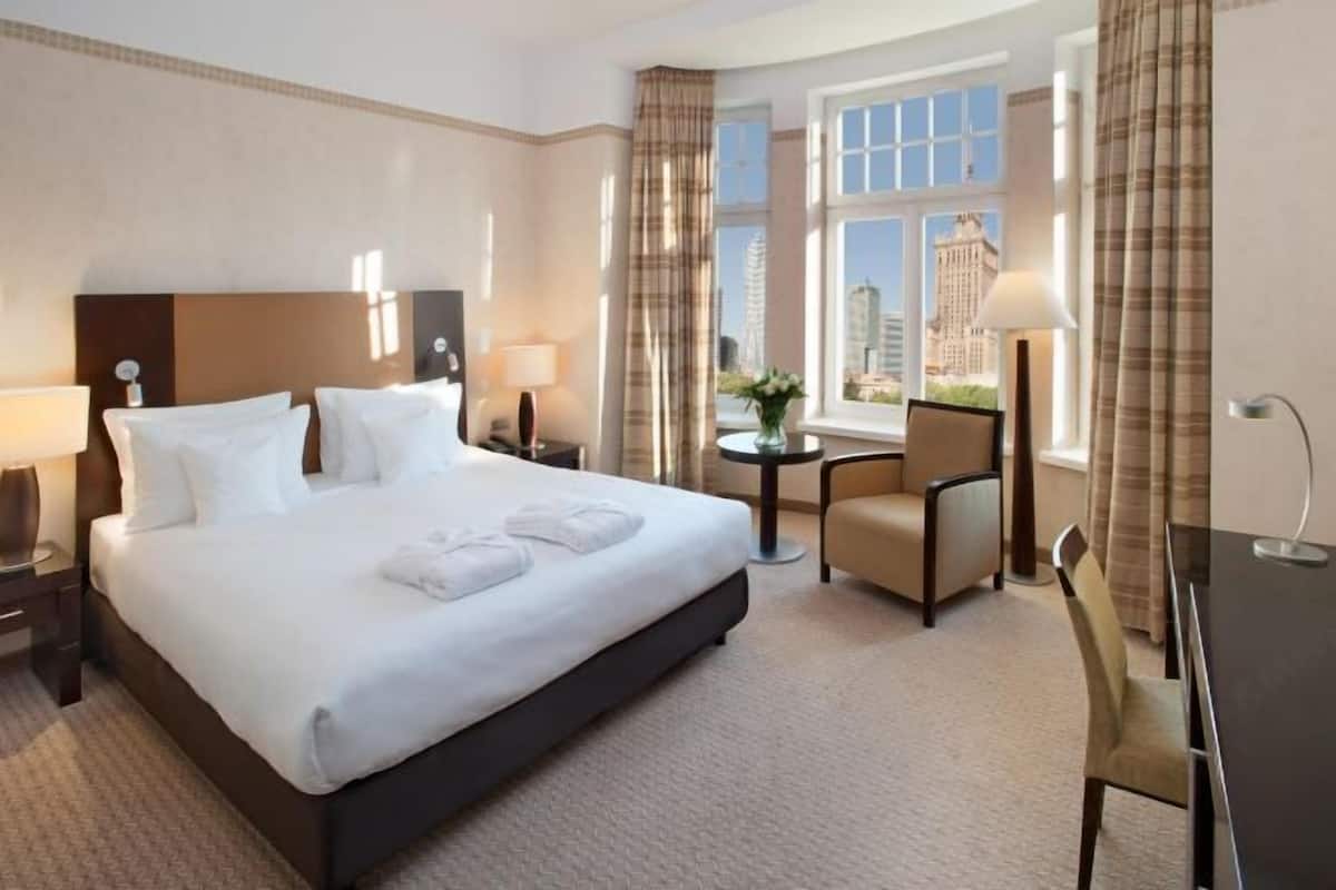 A hotel room with a large bed, bedside tables with lamps, a small round table with a chair, and a view of a cityscape through a window.