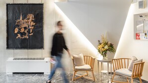 A modern lobby with a marble floor, a large abstract artwork, wooden chairs, a small table with a vase of flowers, and a white air conditioning unit. - Hotel El Greco (Thessaloniki)